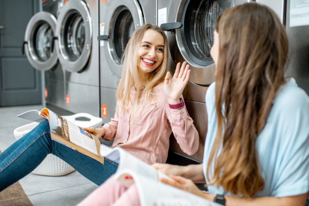 Two cheerful girlfriends having fun together while waiting for the clothes washing in the self-service laundry