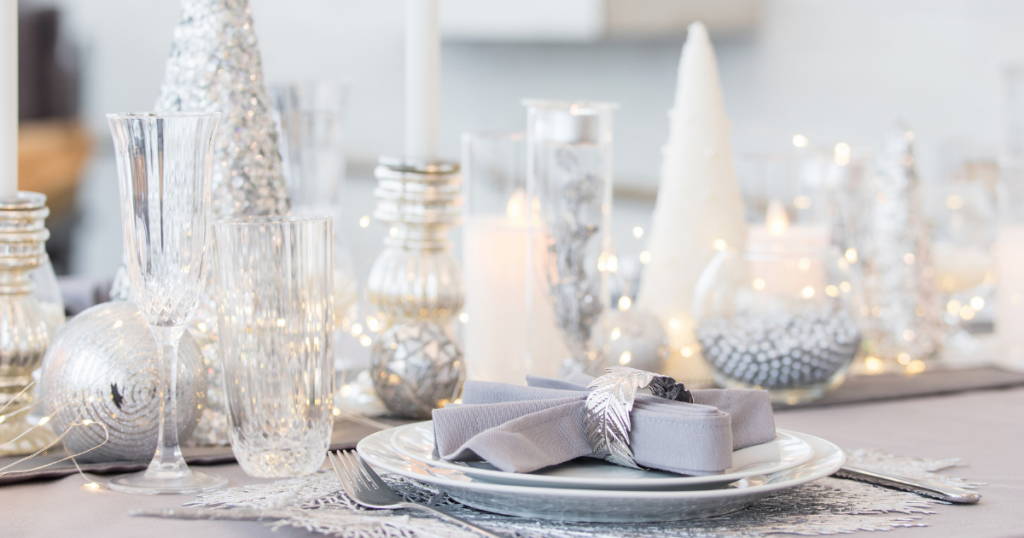 Dinner table set with festive holiday decor that is silver, gray, and white. There are candles, Christmas tree decorations, and silverware on the table.