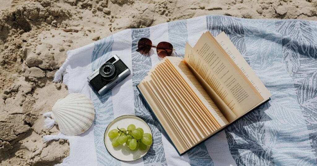A towel on the beach with an assortment of items on it, including a camera, a book, and food.